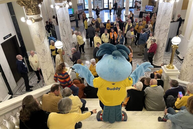South Dakota Public Broadcasting’s mascot, Buddy, waves to a crowd of supporters gathered on Feb. 7, 2025, at the Capitol Rotunda in Pierre. South Dakota Searchlight photo by Seth Tupper.