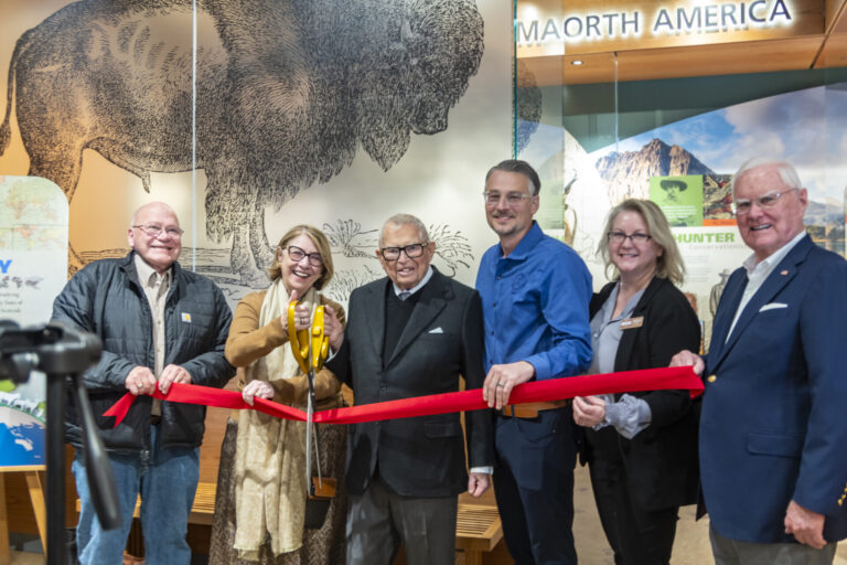 Rosemarie Buntrock cuts a ribbon at the Dacotah Prairie Museum on Tuesday, Dec. 9 as the Rosemarie and Dean L. Buntrock Gallery opens. Also pictured, from left, are Duane Sutton, chairman of the Brown County Commission; Columbia native Dean Buntrock; Aberdeen Mayor Travis Schaunaman; Patricia Kendall, director of the museum; and Terry Birck, project manager for installation of the exhibit. Aberdeen Insider photo by Annie Scott.
