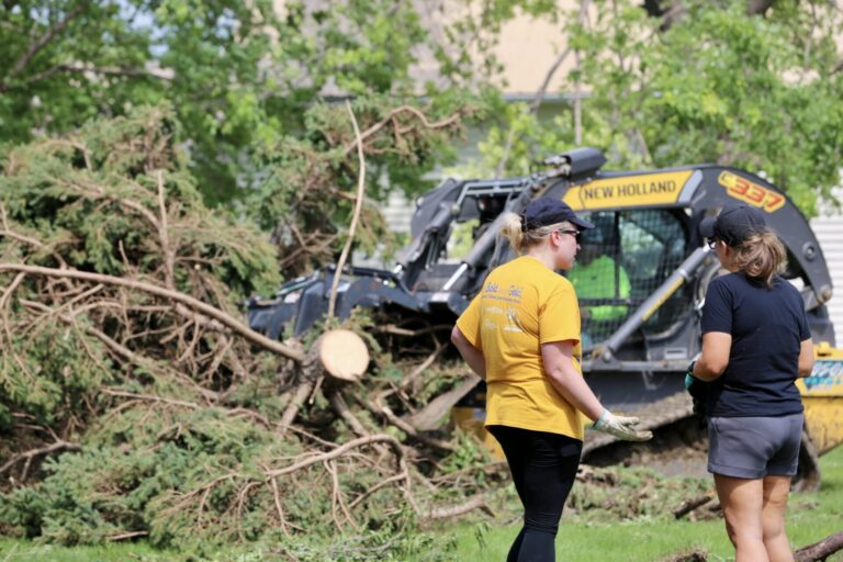 Matilyn Kerr, left, and sister Brittany Kerr watch as a skid-steer loader pulls a tree from Brittany Kerr’s yard in Highmore, South Dakota, on June 29, 2026, after a severe storm that morning. Meghan O’Brien photo courtesy of South Dakota Searchlight.